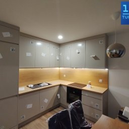 Premium Mebel Dominik Kisielewski - Jasna, narożna kuchnia z drewnianym blatem i oświetleniem LED pod szafkami. Widoczne naklejki na frontach szafek, srebrna lampa wisząca i elementy wyposażenia.