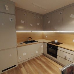 Premium Mebel Dominik Kisielewski - Jasna kuchnia na wymiar z drewnianym blatem i oświetleniem LED pod szafkami. Na frontach szafek widoczne naklejki z kodami. Ujęcie z niskiego kąta.