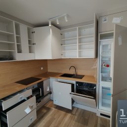 Premium Mebel Dominik Kisielewski - Jasna kuchnia na wymiar z drewnianym blatem w Elblągu. Otwarta zmywarka i szuflady, widoczne półki w szafkach. Nowoczesny design, minimalistyczne oświetlenie.