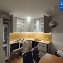 Premium Mebel Dominik Kisielewski - Jasna kuchnia z otwartymi szafkami i sprzętami AGD w trakcie montażu. Widoczne elementy wykończeniowe i oświetlenie podszafkowe. Uwagę zwraca srebrna lampa.