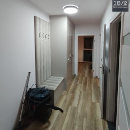 Premium Mebel - Nowoczesny przedpokój z zabudową: szafa, siedzisko z wieszakami. Podłoga imitująca drewno, widoczny odkurzacz. Oświetlenie sufitowe LED.