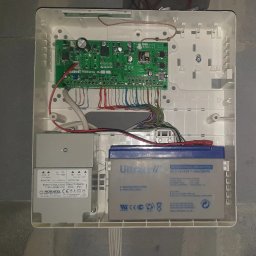 WINKAM - Montaż Monitoringu, Alarmy, Wideodomofony, Smart Home, Instalacja Kamer - Bartosz Gruszka Rybnik - Otwarta obudowa centrali alarmowej z widoczną elektroniką, akumulatorem i transformatorem, zamontowana na ścianie z bloczków. Widoczne przewody i elementy montażowe.