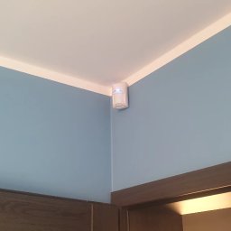 WINKAM - Montaż Monitoringu, Alarmy, Wideodomofony, Smart Home, Instalacja Kamer - Bartosz Gruszka Rybnik - Biały czujnik ruchu z niebieską diodą LED zamontowany w rogu pokoju o błękitnych ścianach i białym suficie, nad ciemnobrązową framugą drzwi.