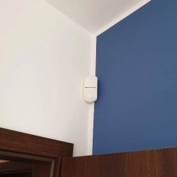 WINKAM - Montaż Monitoringu, Alarmy, Wideodomofony, Smart Home, Instalacja Kamer - Bartosz Gruszka Rybnik - Biały czujnik ruchu zamontowany w rogu pokoju, na styku białej i niebieskiej ściany, z widocznym fragmentem drewnianej ramy drzwiowej. Minimalistyczny design.