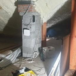 WINKAM - Montaż Monitoringu, Alarmy, Wideodomofony, Smart Home, Instalacja Kamer - Bartosz Gruszka Rybnik - Montaż punktu dostępowego Wi-Fi na ceglanym kominie w surowym wnętrzu poddasza z izolacją piankową. Widoczne kable, narzędzia i materiały instalacyjne.