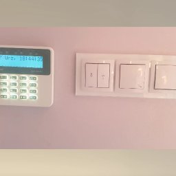 WINKAM - Montaż Monitoringu, Alarmy, Wideodomofony, Smart Home, Instalacja Kamer - Bartosz Gruszka Rybnik - Panel alarmowy Satel Perfecto z wyświetlaczem i klawiaturą numeryczną, zamontowany obok białych, szklanych włączników światła na jasnoróżowej ścianie.