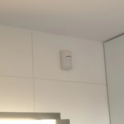 WINKAM - Montaż Monitoringu, Alarmy, Wideodomofony, Smart Home, Instalacja Kamer - Bartosz Gruszka Rybnik - Zamontowany czujnik ruchu alarmu w rogu pomieszczenia, na białej ścianie z widocznymi fugami. Obok fragment białej szafki. Minimalistyczny design.