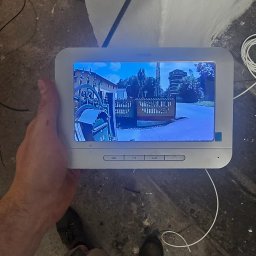 WINKAM - Montaż Monitoringu, Alarmy, Wideodomofony, Smart Home, Instalacja Kamer - Bartosz Gruszka Rybnik - Ekran wideodomofonu HiLook w dłoni, wyświetlający obraz z kamery na zewnątrz. Widoczne przewody, kombinerki i fragment buta. Szary beton na podłodze.