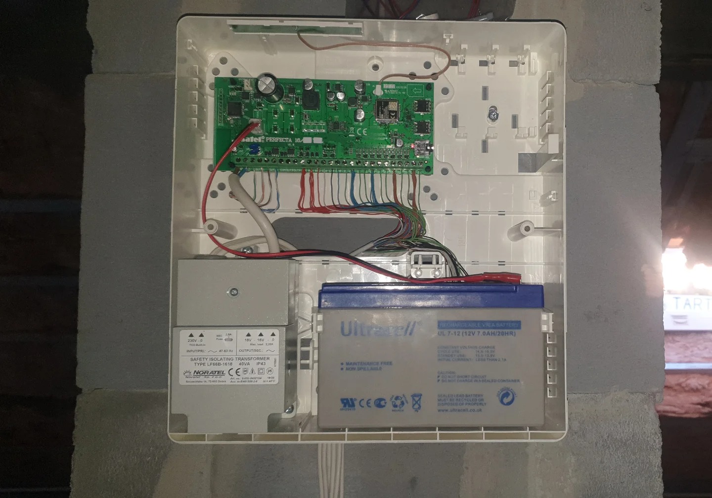 Otwarta obudowa centrali alarmowej z widoczną elektroniką, akumulatorem i transformatorem, zamontowana na ścianie z bloczków. Widoczne przewody i elementy montażowe.