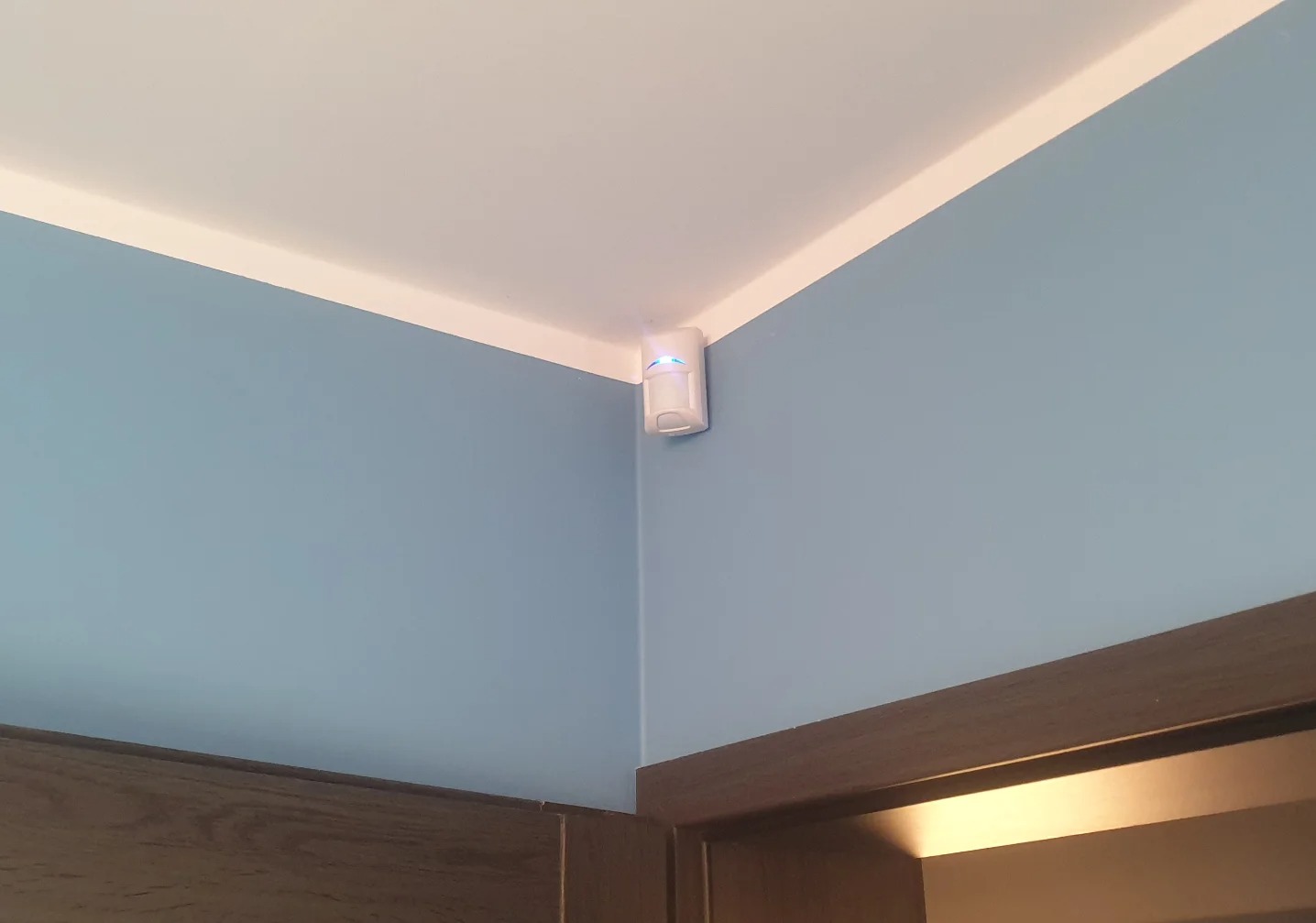Biały czujnik ruchu z niebieską diodą LED zamontowany w rogu pokoju o błękitnych ścianach i białym suficie, nad ciemnobrązową framugą drzwi.