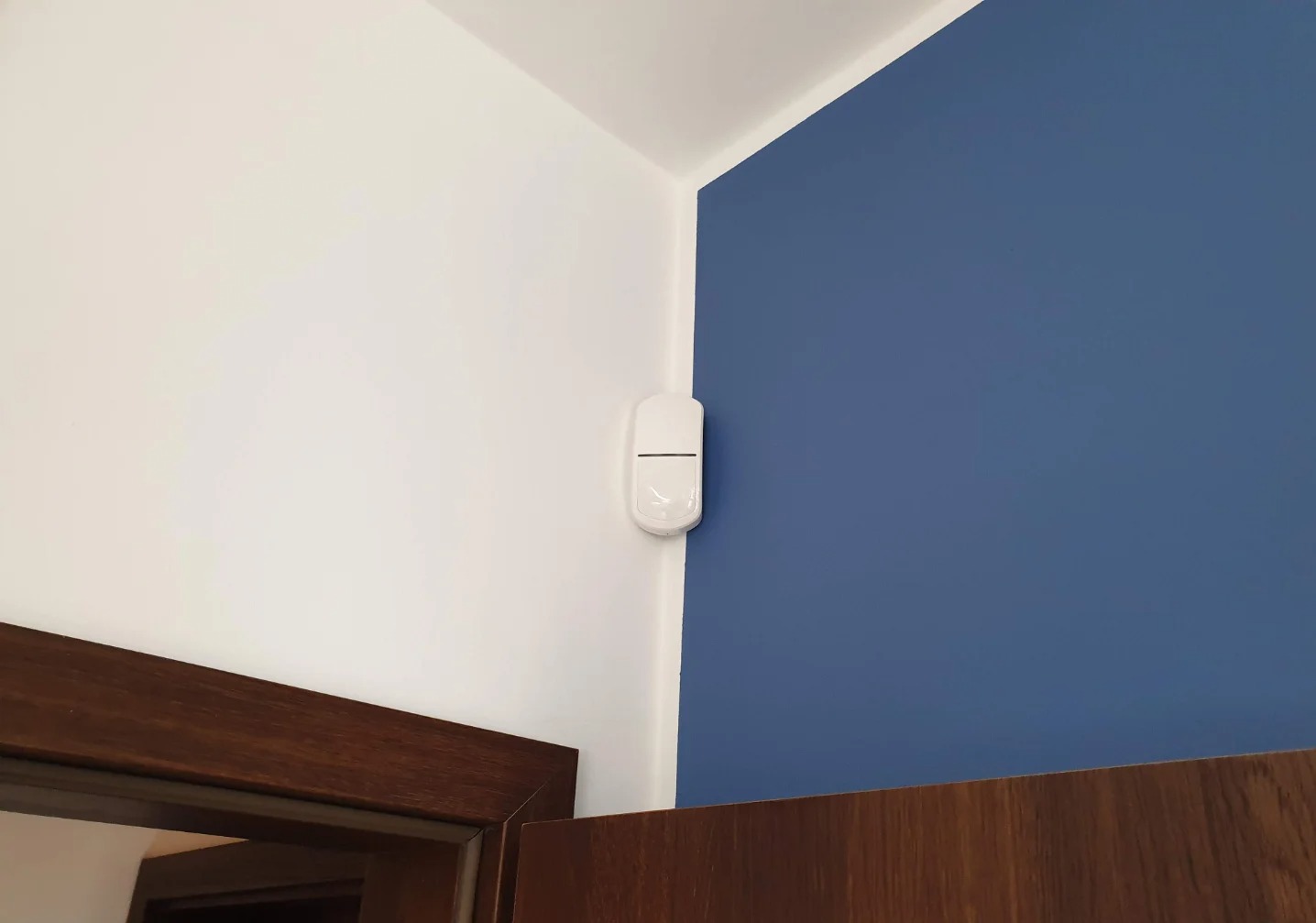Biały czujnik ruchu zamontowany w rogu pokoju, na styku białej i niebieskiej ściany, z widocznym fragmentem drewnianej ramy drzwiowej. Minimalistyczny design.