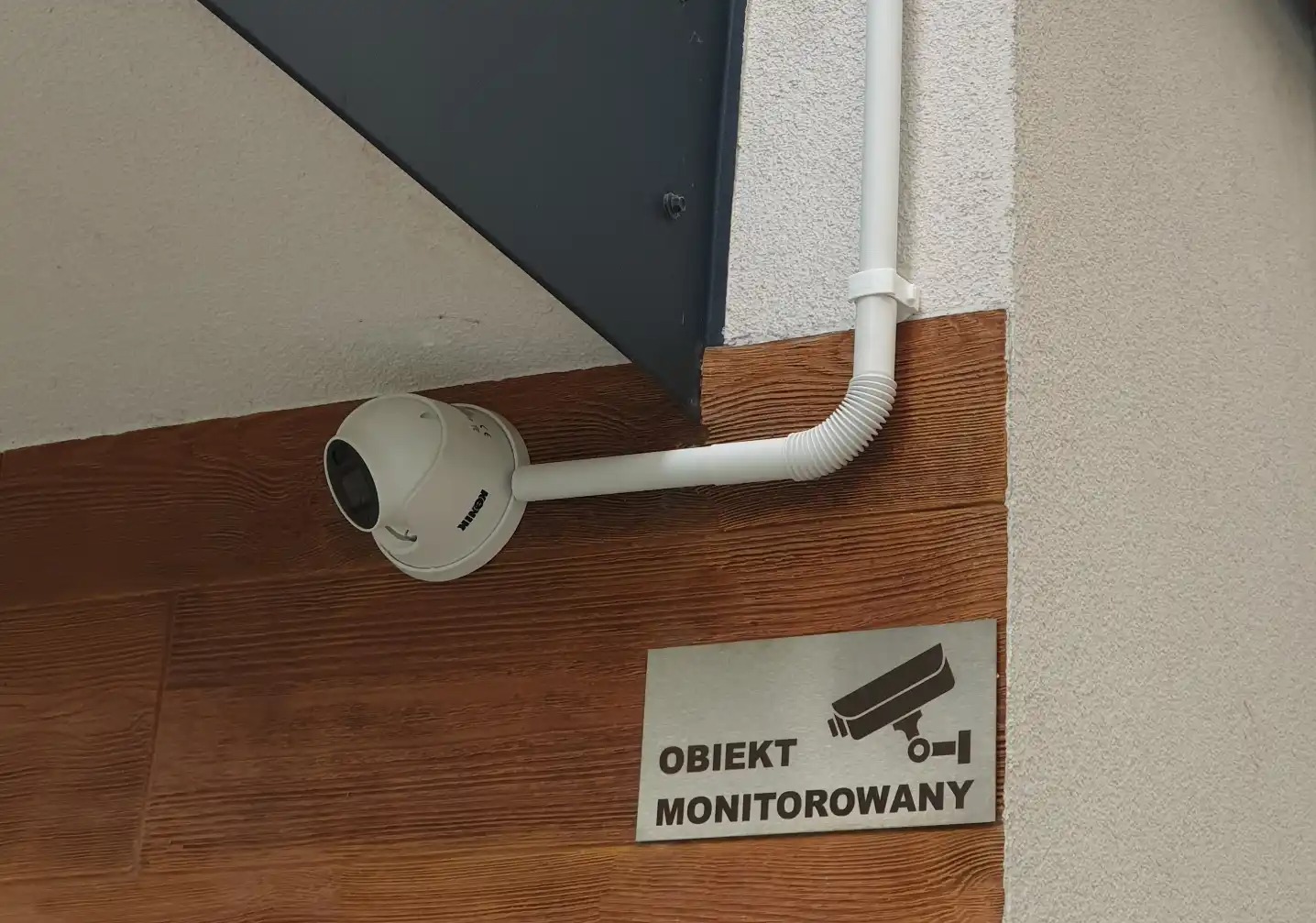 Kamera monitoringu kopułkowa marki Konik, zamontowana na ścianie z drewnopodobną okładziną, obok tabliczka 'Obiekt Monitorowany'.