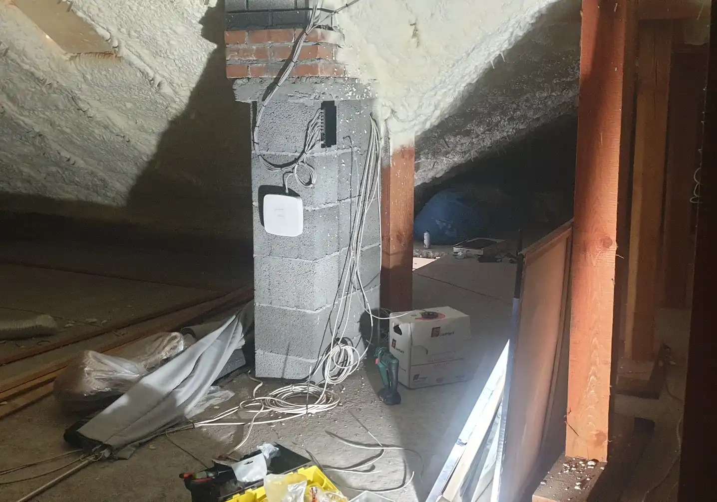 Montaż punktu dostępowego Wi-Fi na ceglanym kominie w surowym wnętrzu poddasza z izolacją piankową. Widoczne kable, narzędzia i materiały instalacyjne.