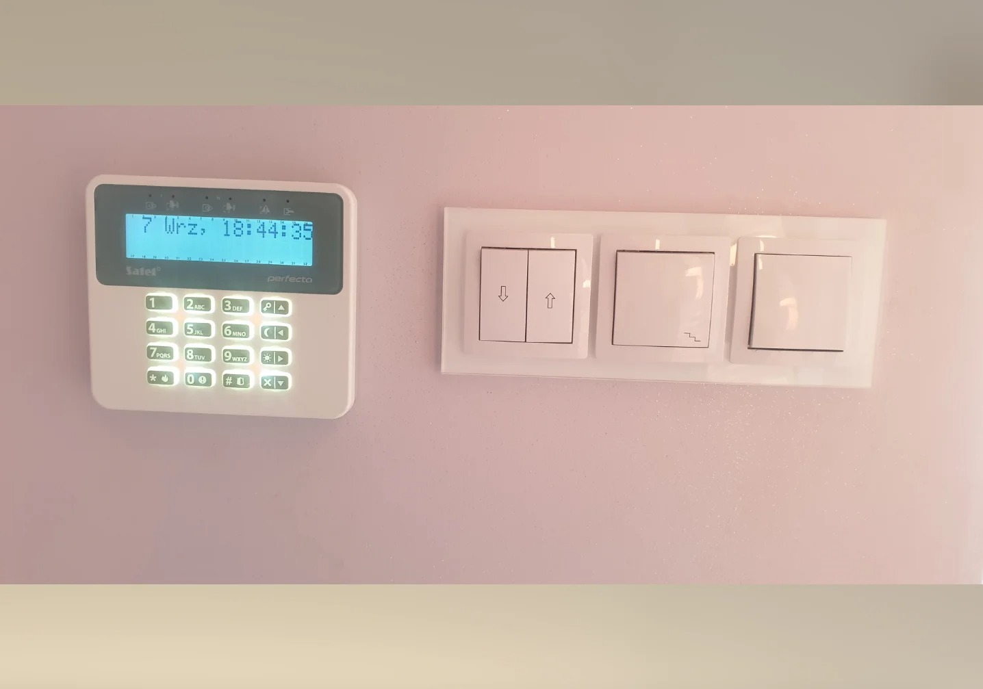 Panel alarmowy Satel Perfecto z wyświetlaczem i klawiaturą numeryczną, zamontowany obok białych, szklanych włączników światła na jasnoróżowej ścianie.