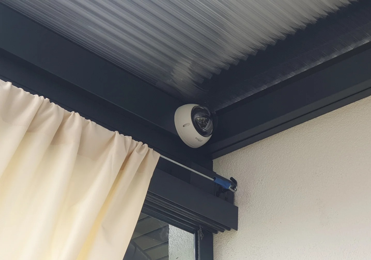 Kamera monitoringu IP zamontowana pod zadaszeniem tarasu, widoczna kremowa zasłona i fragment okna. Biała ściana i szara konstrukcja zadaszenia.