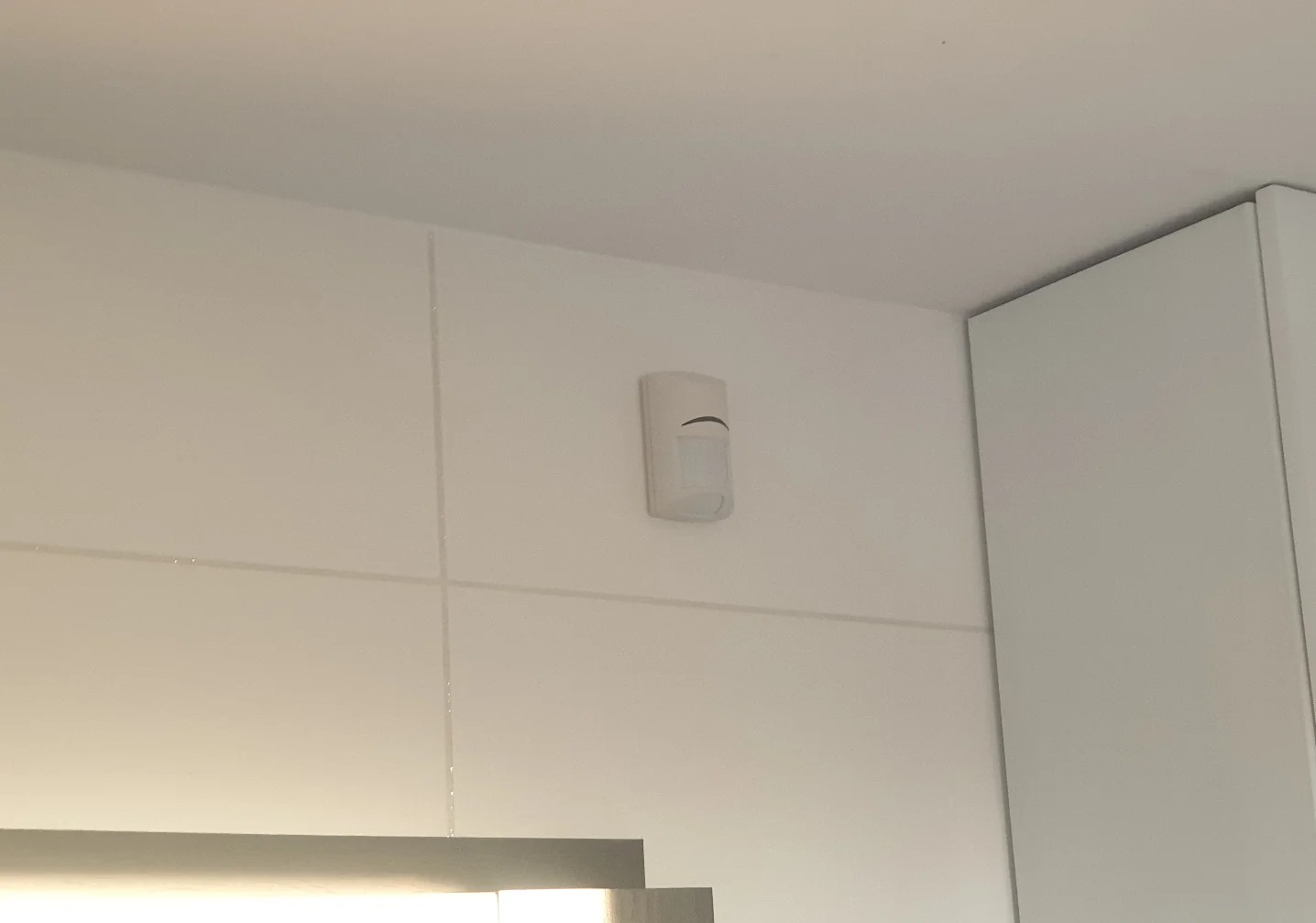 Zamontowany czujnik ruchu alarmu w rogu pomieszczenia, na białej ścianie z widocznymi fugami. Obok fragment białej szafki. Minimalistyczny design.