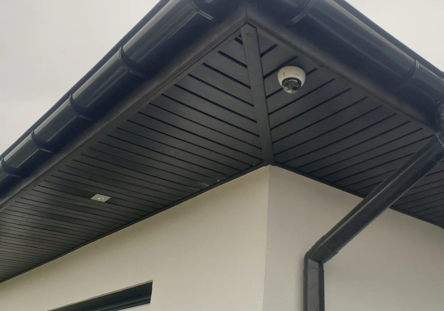 Kamera monitoringu zamontowana pod okapem budynku z czarną podbitką. Widoczny fragment rynny i oświetlenie punktowe.