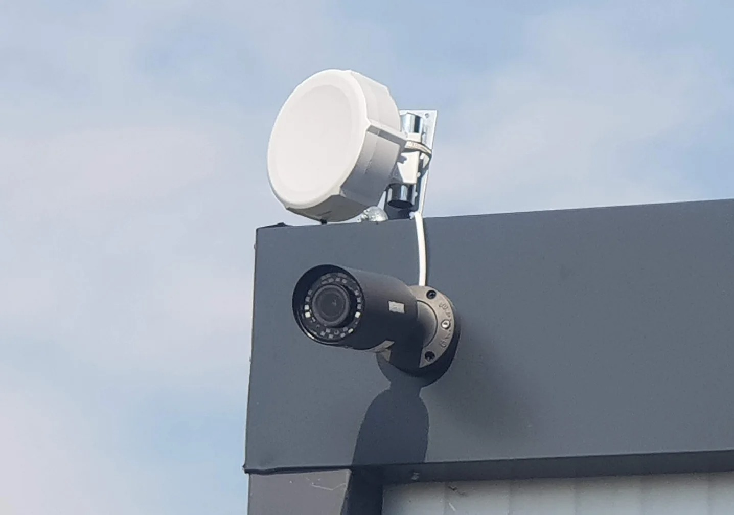 Zewnętrzna kamera monitoringu CCTV z anteną Wi-Fi na szarym budynku, skierowana w dół, widok z dołu na błękitne niebo. Zabezpieczenia domu i firmy.
