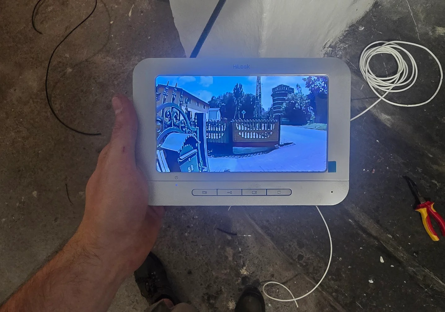 Ekran wideodomofonu HiLook w dłoni, wyświetlający obraz z kamery na zewnątrz. Widoczne przewody, kombinerki i fragment buta. Szary beton na podłodze.
