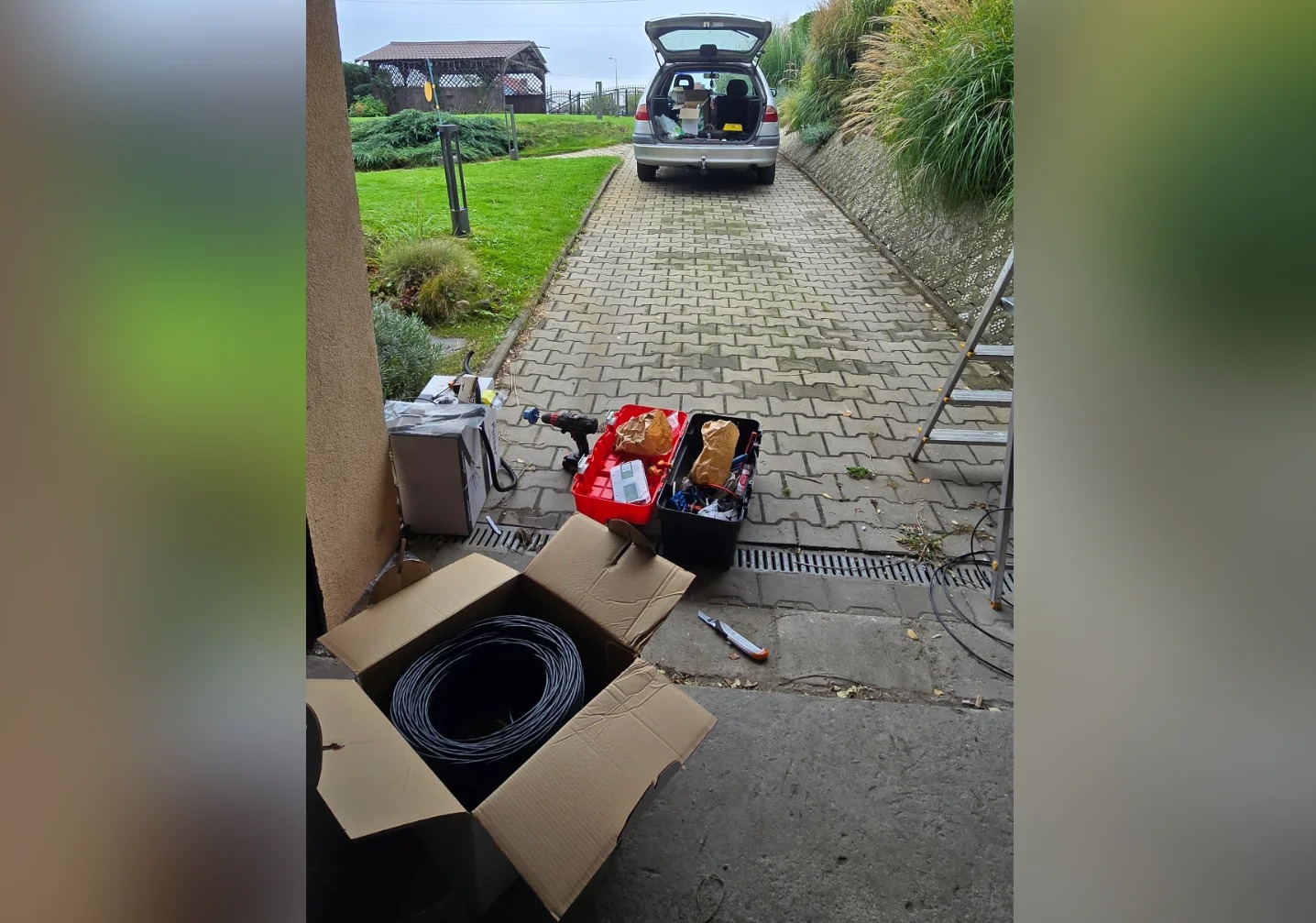 Rozłożone narzędzia i kable elektryczne przed domem z otwartym bagażnikiem samochodu, sugerujące przygotowania do instalacji elektrycznej na zewnątrz budynku.