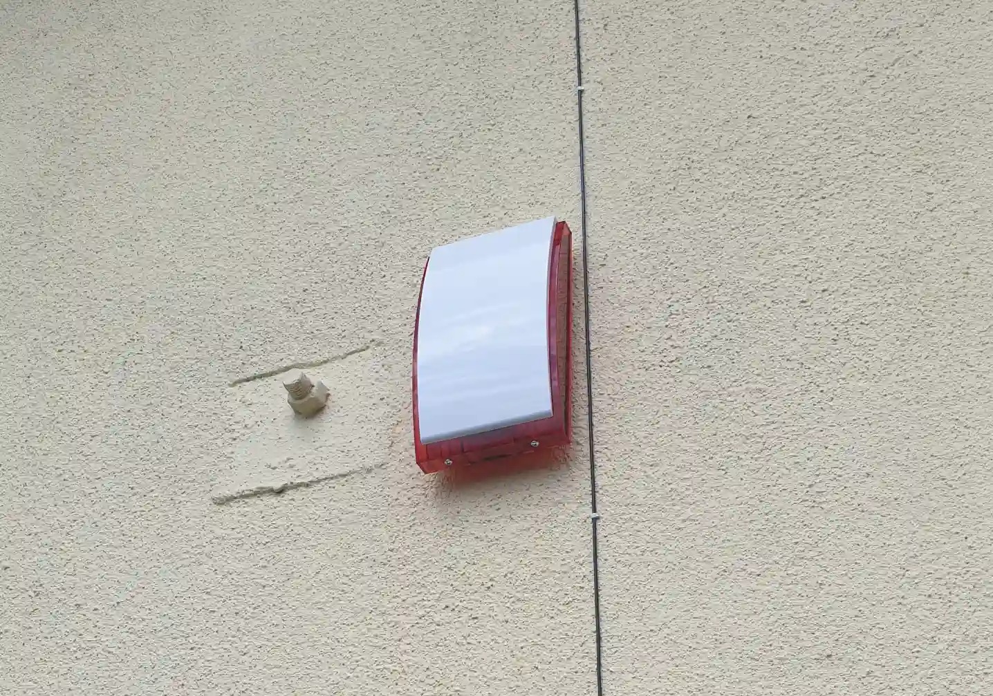 Zewnętrzna sygnalizacja alarmowa z biało-czerwoną obudową, zamontowana na teksturowanej elewacji budynku. Obok widoczny uchwyt montażowy i przewód.