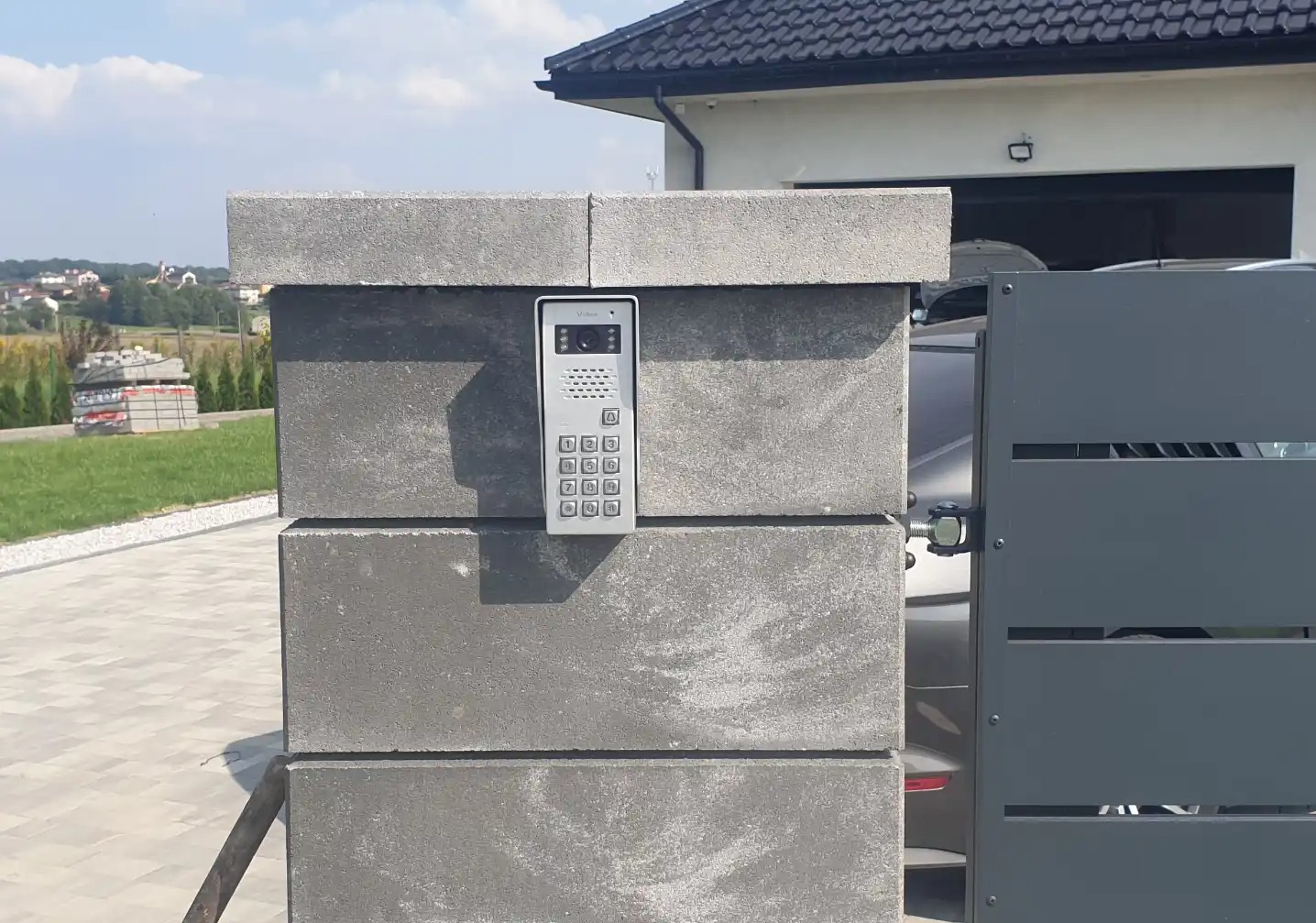 Nowoczesny wideodomofon Vidos zamontowany na słupku z szarej kostki betonowej przy metalowej bramie posesji. Widok na okolicę w tle.