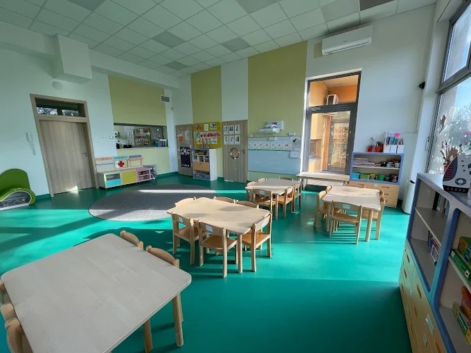 Jasne wnętrze sali zabaw z drewnianymi stolikami i krzesłami, zieloną podłogą i kolorowymi szafkami. Widoczne okno z wpadającym światłem dziennym i tablica.