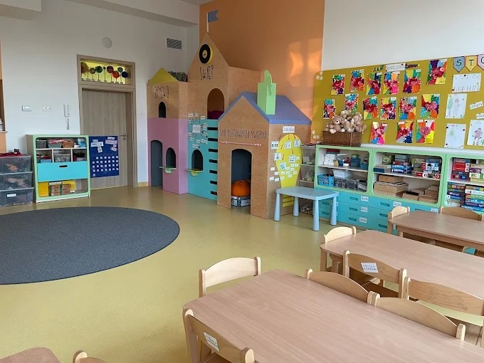 Jasne wnętrze sali zabaw z drewnianą zabudową w kształcie miasteczka, stolikami i krzesłami. Na ścianach wiszą dziecięce prace plastyczne i edukacyjne pomoce.