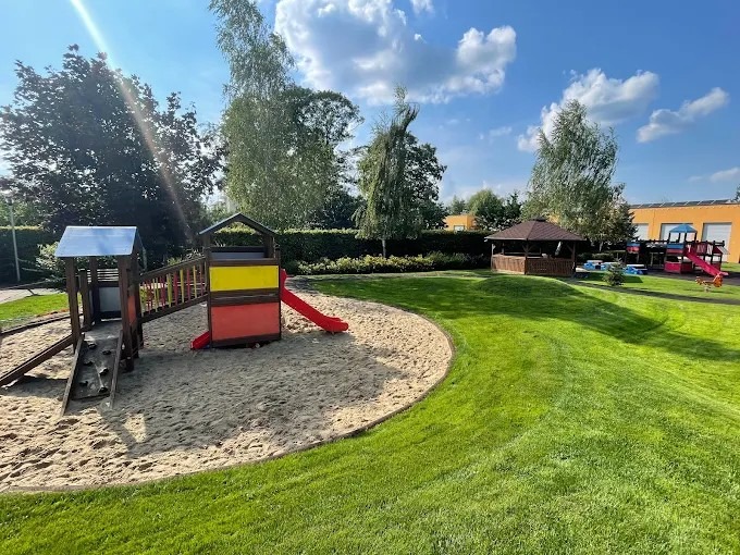 Kolorowy plac zabaw z piaskownicą i zjeżdżalnią na zielonym terenie żłobka, w tle altana i budynek z panelami słonecznymi. Słoneczny dzień.