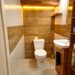 PMP Paweł Paciora - Nowoczesna łazienka z drewnianymi akcentami: szafka nad toaletą z oświetleniem LED, umywalka z szafką, płytki imitujące drewno, białe płytki i jasna podłoga.