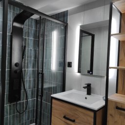 PMP Paweł Paciora - Nowoczesna łazienka z kabiną prysznicową typu walk-in, czarną armaturą i szafką z drewnianym frontem. Podświetlane lustro nad umywalką. Ciemnozielone płytki na ścianie.