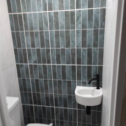 PMP Paweł Paciora - Wykończona toaleta: pionowe, grafitowe płytki na ścianie, biała umywalka z czarną armaturą i toaleta. Minimalistyczny design, wąskie pomieszczenie.