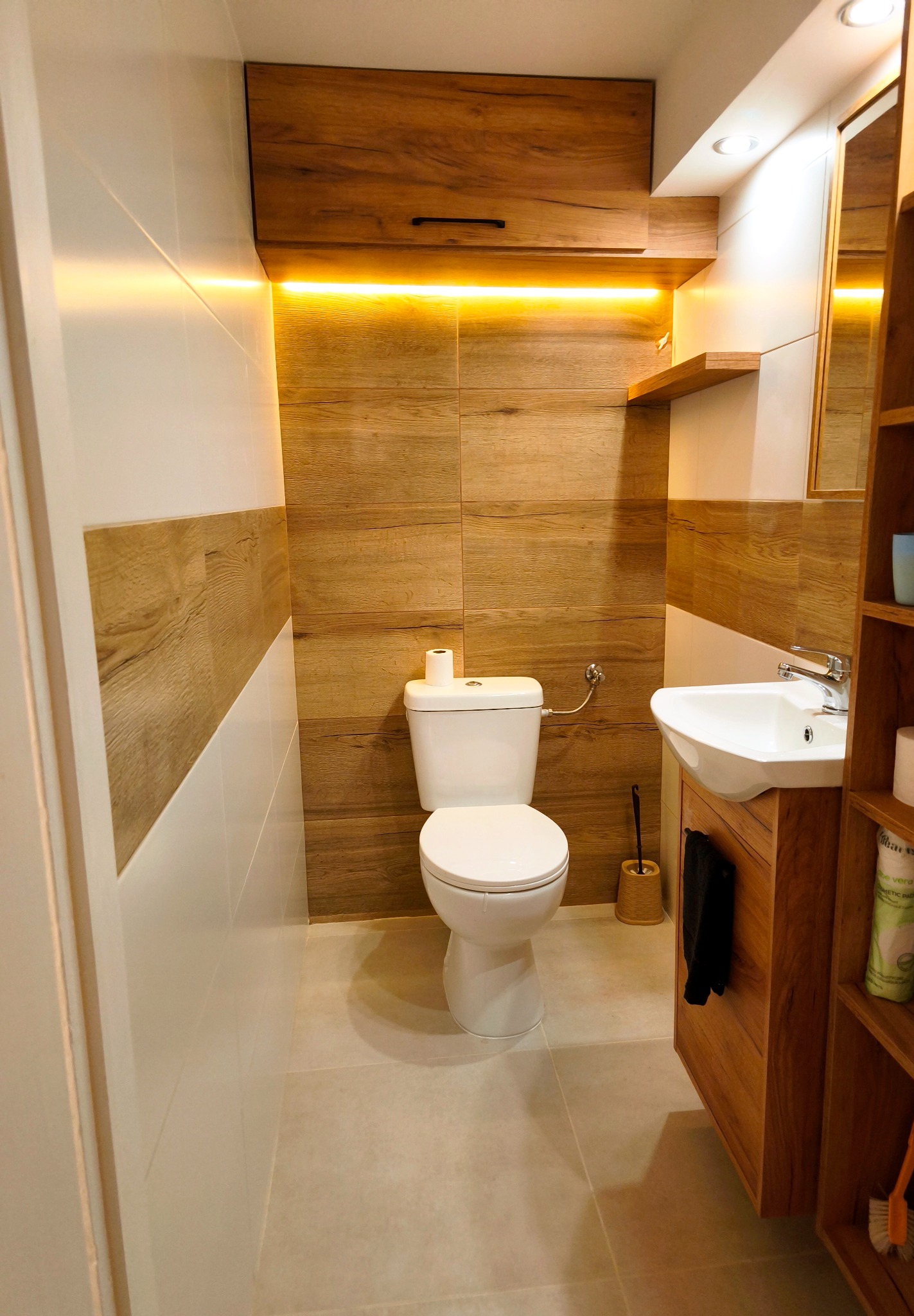 Nowoczesna łazienka z drewnianymi akcentami: szafka nad toaletą z oświetleniem LED, umywalka z szafką, płytki imitujące drewno, białe płytki i jasna podłoga.