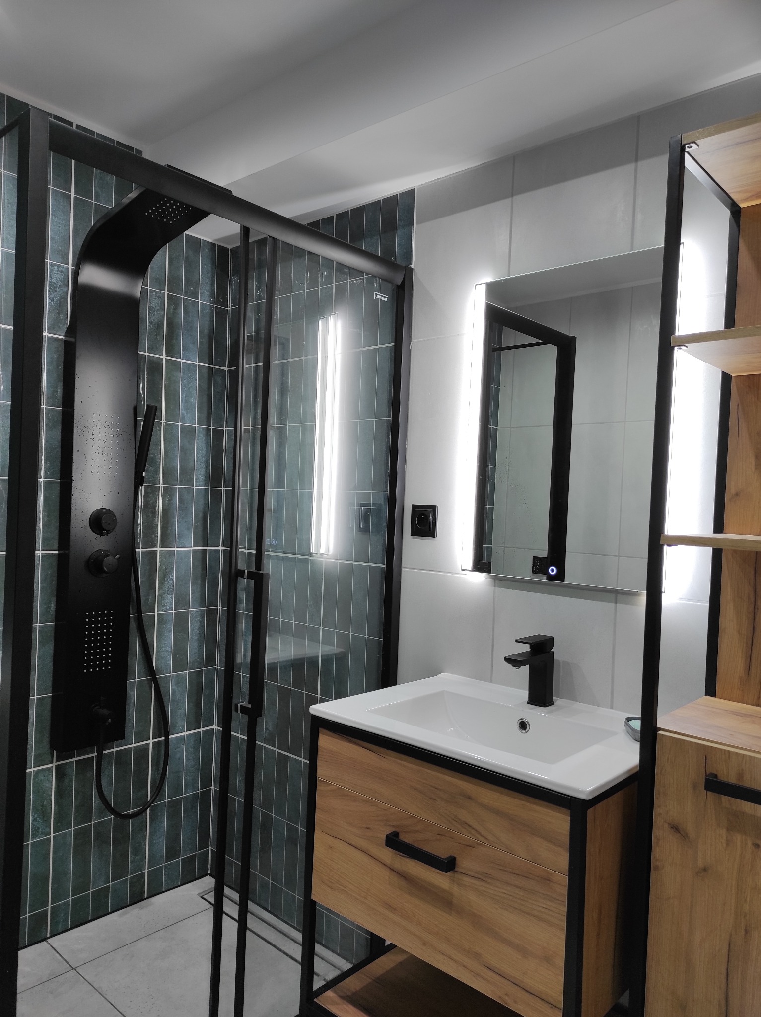 Nowoczesna łazienka z kabiną prysznicową typu walk-in, czarną armaturą i szafką z drewnianym frontem. Podświetlane lustro nad umywalką. Ciemnozielone płytki na ścianie.