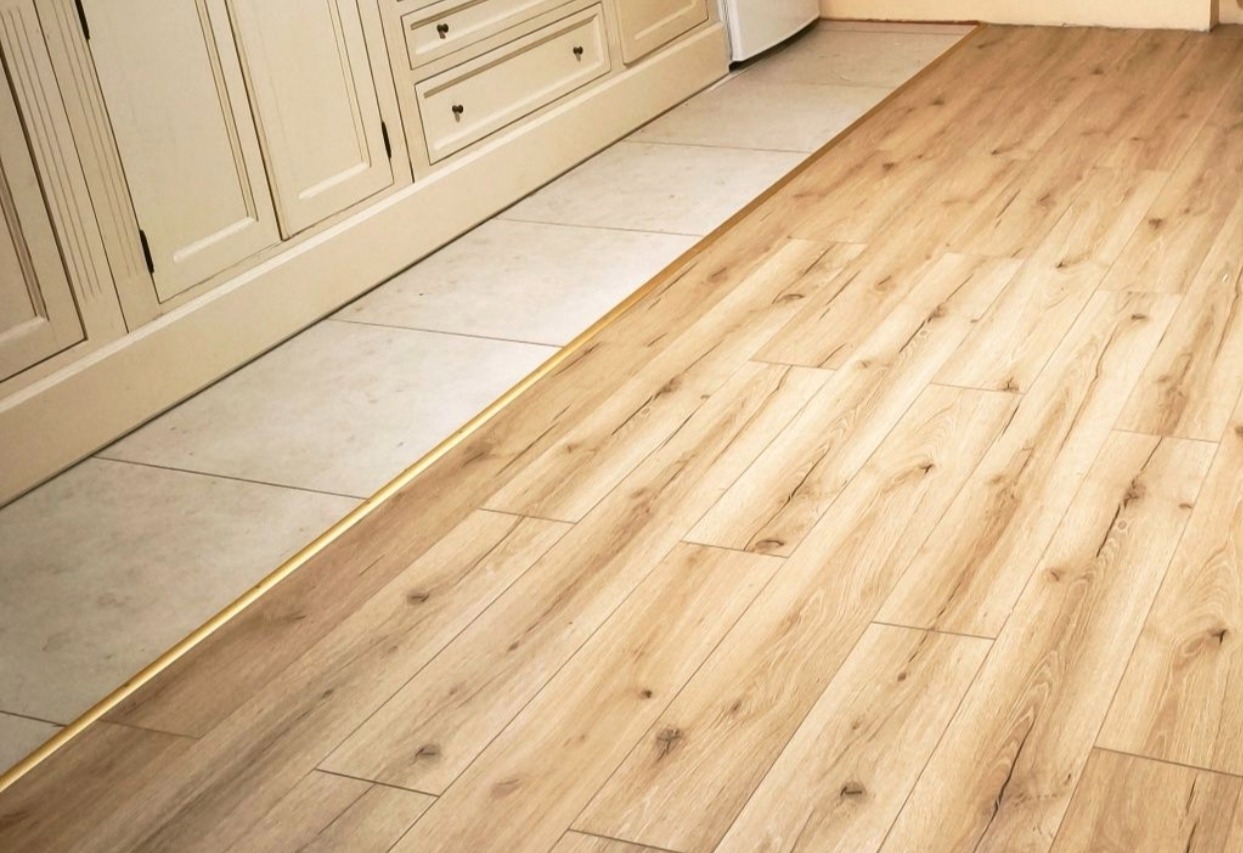 Jasne panele podłogowe ułożone wzdłuż szafki kuchennej, oddzielone złotą listwą od jasnych płytek. Widok z góry, ukazujący estetyczne połączenie materiałów.