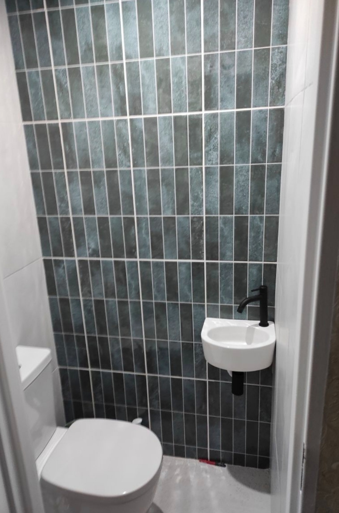 Wykończona toaleta: pionowe, grafitowe płytki na ścianie, biała umywalka z czarną armaturą i toaleta. Minimalistyczny design, wąskie pomieszczenie.