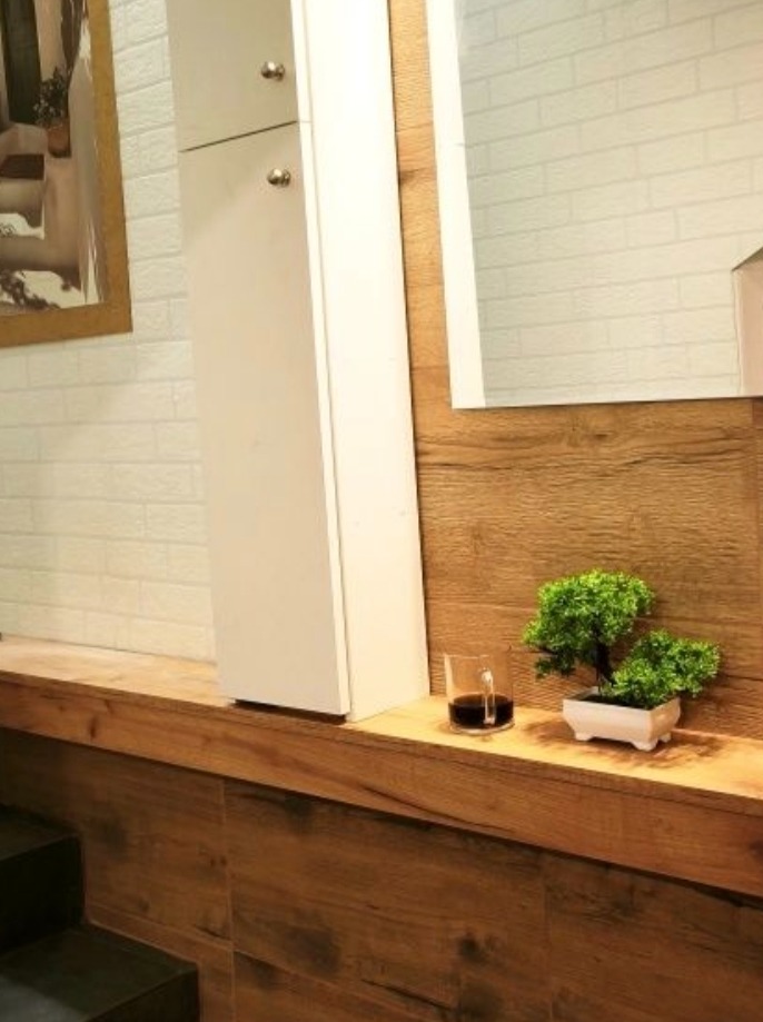 Nowoczesna łazienka z białą szafką, drewnianym blatem, ceglaną ścianą i dekoracjami: szklanka z napojem i miniaturowe drzewko bonsai w białej doniczce.