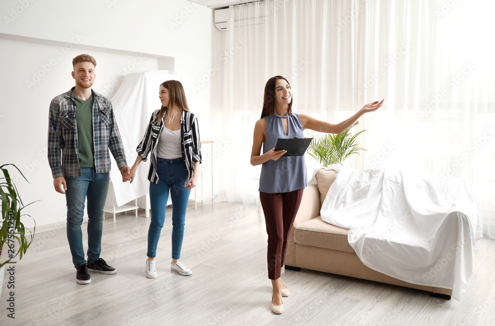 Agentka nieruchomości prezentuje jasne, przestronne wnętrze młodemu małżeństwu. Sofa przykryta białym płótnem, duże okno z firankami, roślina doniczkowa.