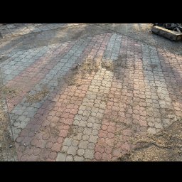 Doktor Bruk - Ułożona kostka brukowa w odcieniach szarości i czerwieni, pokryta piaskiem i drobnymi zabrudzeniami. Widoczne nierówności i ślady użytkowania.