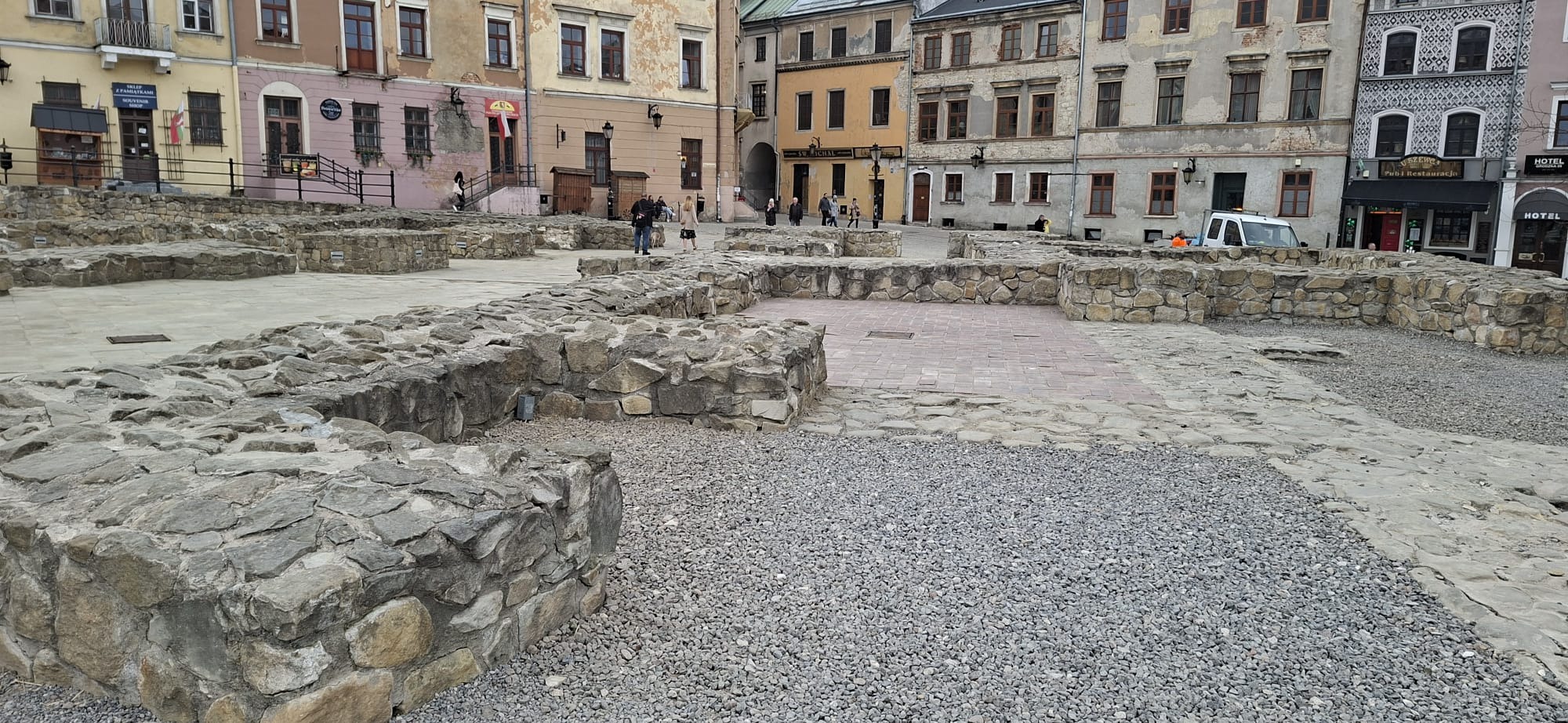 Odkryte fundamenty budynku na placu miejskim, otoczone żwirem i kamiennymi murkami. W tle zabudowa miejska z różnorodną architekturą i przechodzący ludzie.