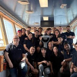 NeoKoszulki.pl - hurtowania odzieży reklamowej i roboczej - Grupowe zdjęcie załogi w ciemnych koszulkach z logo firmy, stojącej w sterowni statku, z widokiem na morze za oknami. Uśmiechnięci ludzie w jednakowych strojach.