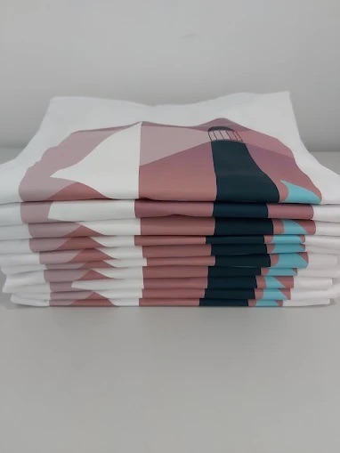 Stos białych koszulek z nadrukiem w geometryczny wzór latarni morskiej, wykonanym w odcieniach różu, błękitu i granatu. Realizacja z Warszawy.