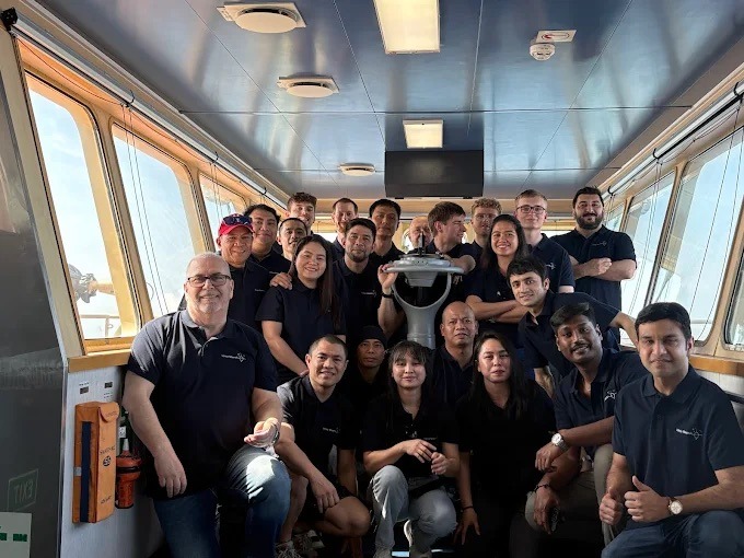 Grupowe zdjęcie załogi w ciemnych koszulkach z logo firmy, stojącej w sterowni statku, z widokiem na morze za oknami. Uśmiechnięci ludzie w jednakowych strojach.