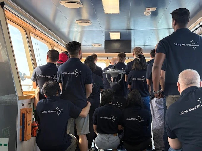 Grupa osób w jednakowych, granatowych koszulkach polo z logo Vilnia Maersk, stojących w jasnym wnętrzu przypominającym kabinę statku. Widok z tyłu, skupienie na nadruku.