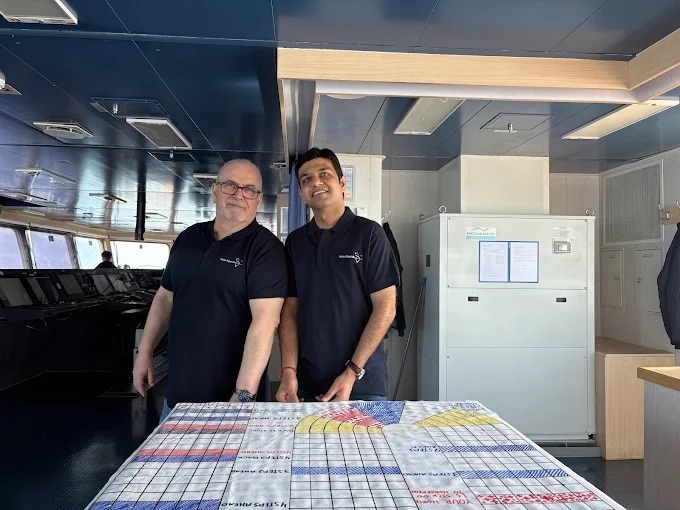 Dwaj mężczyźni w czarnych koszulkach polo z logo firmy stoją za stołem z nadrukiem w pomieszczeniu przypominającym mostek kapitański statku.