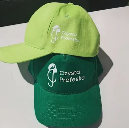 Dwie czapki z daszkiem: limonowa i zielona, z białym nadrukiem logo 'Czysta Profeska'. Widoczne detale szwów i materiału. Ułożone jedna na drugiej.