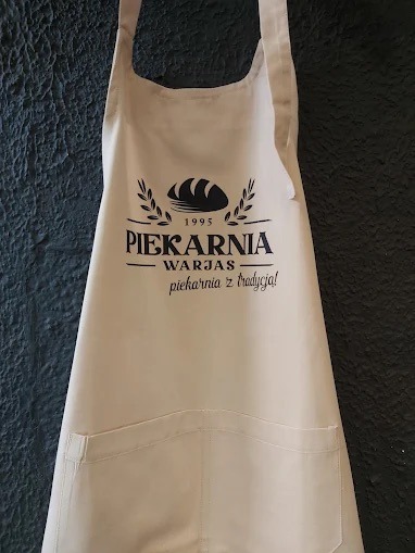 Beżowy fartuch z czarnym nadrukiem 'Piekarnia Warjas 1995 - piekarnia z tradycją!', wiszący na ciemnym tle. Widoczne kieszenie i szelki.