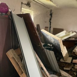 Czyściuś-Trans - Stare drzwi, palety i gruz w zrujnowanym pomieszczeniu z pajęczynami. Widoczne ślady wilgoci na ścianach i suficie. Chaos i zaniedbanie.