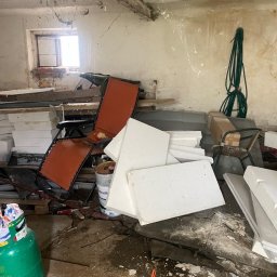 Czyściuś-Trans - Zagracone pomieszczenie z oknem, wypełnione styropianem, starymi meblami i narzędziami. Widoczny bałagan i pozostałości po pracach budowlanych.