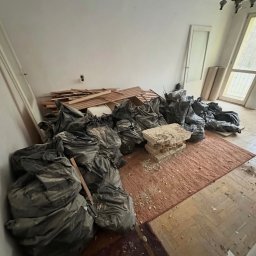 Czyściuś-Trans - Pokój przed opróżnianiem: worki na śmieci, gruz, deski na dywanie i parkiecie. Widoczne okno i drzwi. Ujęcie z góry, oświetlenie naturalne.